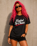A woman with bright pink hair and sunglasses wears a black graphic T-shirt with the text 'Fueled By Chaos' in white script and a red underline, paired with black denim shorts, standing against a concrete wall.