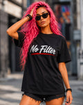 A woman with bright pink hair and black sunglasses wears a black graphic T-shirt with the words 'No Filter' in white script and a red underline, paired with black denim shorts, standing on a blurred city street.